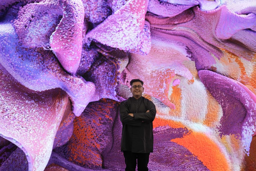 The media artist Refik Anadol standing with his arms crossed in front of a huge, fluid data sculpture in purple and pink created by artificial intelligence.